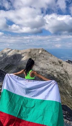 a woman holding a large flag on top of a mountain with mountains in the background