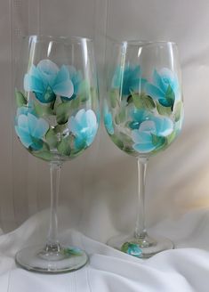 two wine glasses with blue flowers painted on the glass are sitting on a white sheet