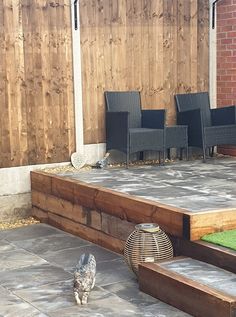 a cat walking across a patio next to some chairs and table with grass on the ground