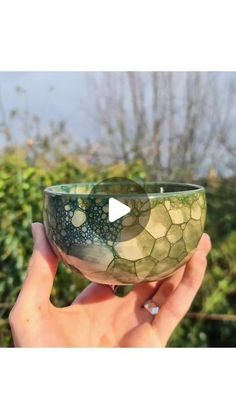 a hand holding a green and white bowl in it's left hand, with trees in the background