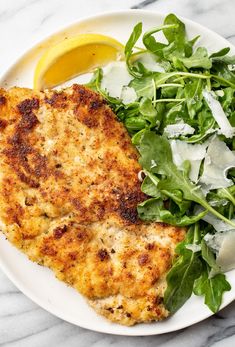 a white plate topped with chicken next to a green salad and lemon wedge on top of a marble table