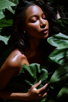 a woman with long black hair is surrounded by green leaves and has her eyes closed