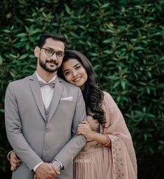 a man and woman standing next to each other in front of some bushes with their arms around each other