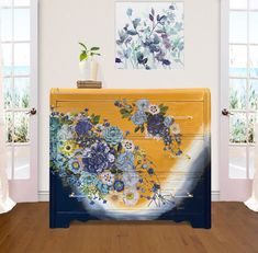 a yellow and blue chest with flowers painted on it in front of two open windows