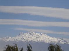 a snow covered mountain in the distance with trees around it and blue sky behind it