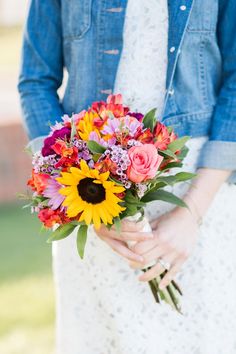 a woman holding a bouquet of colorful flowers in her hands and wearing a denim jacket