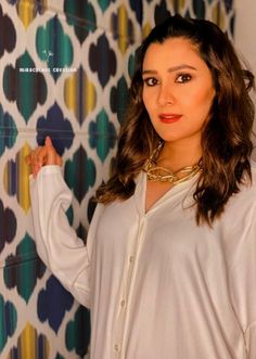 a woman standing in front of a wall with colorful tiles on it and wearing a white shirt