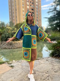 a woman standing on top of a rock next to a body of water wearing a green and blue crocheted coat