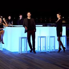 a group of people standing around a bar with stools and lights on the floor