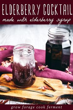 a wooden cutting board topped with a mason jar filled with elderberry cocktail syrup and two spoons