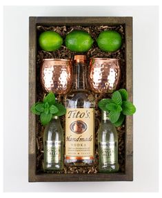 a wooden box with three bottles and two glasses filled with limes, minty garnish