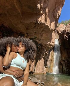 two women sitting next to each other in front of a waterfall and rock formation with water running down it