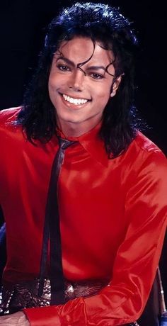 a man with long black hair wearing a red shirt and tie smiling at the camera