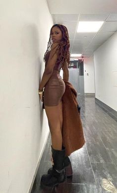 a woman leaning against a wall wearing boots and a brown dress with long hair, standing in an office hallway