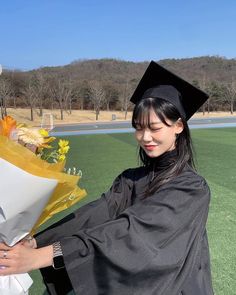 a woman in a graduation gown holding flowers and a bouquet on the side of a field