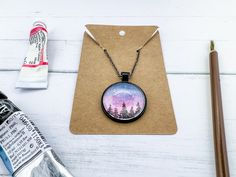 a bottle of paint next to a necklace with trees on it and an acrylic pen