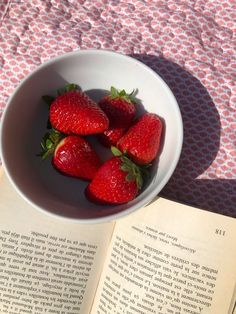 strawberries in a white bowl on top of an open book next to a pink and white tablecloth