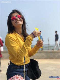 a woman in yellow shirt and sunglasses holding up a small kite with people walking on the beach behind her