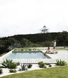 two people sitting on the ground near a swimming pool with plants and trees in the background