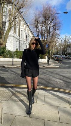 a woman in short shorts and boots is standing on the sidewalk with her hands behind her head