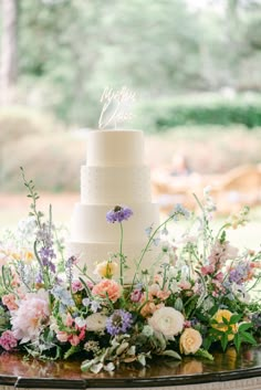 a wedding cake with flowers and greenery on the table in front of an outdoor setting