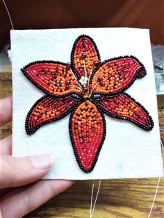 someone is holding up a piece of beaded art with an orange flower on it