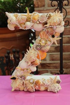 a letter made out of seashells and pearls on a pink table with a potted plant in the background