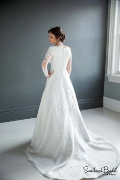 a woman in a white wedding dress standing by a window with her back to the camera