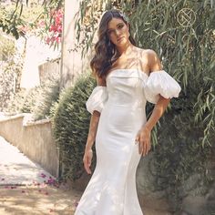 a woman wearing a white dress standing in front of a tree and bushes with her hands on her hips