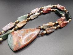 This lovely necklace showcases an exquisite Ocean Jasper pendant in enchanting hues of red, green, gray, and brown. The necklace is elegantly complemented by 10 to 20 mm figural Ocean Jasper beads, along with high-quality misty Quartz faceted beads and Czech fire-polished beads. The necklace is elegantly secured with a gold-plated hook clasp, guaranteeing both durability and style. Necklace length: 26 inches/67 cm. Pendant measures 58 by 35 mm/2,3 in by 1,4 in.  Indulge in the beauty of nature a Elegant Jasper Necklace For Gift, Handmade Multicolor Jasper Jewelry, Jasper Gemstone Beaded Necklaces As Gift, Jasper Gemstone Beaded Necklace As Gift, Unique Multicolor Jasper Necklaces, Multicolor Jasper Jewelry For Gift, Multicolor Jasper Necklace For Gift, Elegant Jasper Beaded Necklace For Gift, Quartz Pendant Necklace