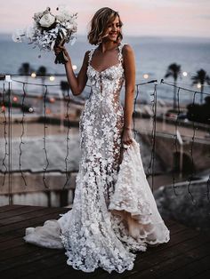 a woman standing on top of a wooden deck next to the ocean holding a bouquet of flowers