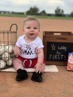 a baby sitting on the ground next to a basket of baseballs and a sign that says we way to first