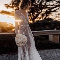 a woman wearing a wedding dress with long sleeves and flowers in her hand is standing outside