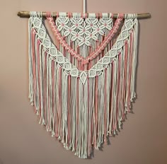 a macrame wall hanging with pink and white yarn on the top, attached to a wooden pole