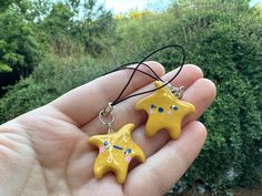 a hand holding two small yellow stars on black cord necklaces in front of some bushes