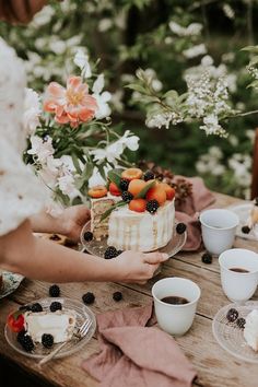 a woman holding a piece of cake on top of a wooden table next to cups and saucers