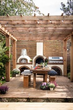 an outdoor kitchen and dining area with wood pergolated roofing, stone patio furniture and brick oven