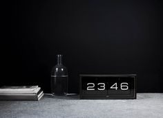 an alarm clock sitting next to a bottle and stack of books on the floor in front of a black wall