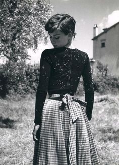 a woman wearing a skirt and black top standing in front of a tree with her hands on her hips