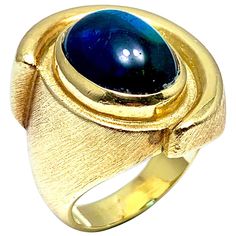 A gorgeous Indicolite Tourmaline fashion ring designed by Bruno Guidi. The 6.43 cabochon Indicolite displays a lustrous teal color, bezel set in an 18 karat yellow gold abstarct design ring. The inside shank is signed "Bruno Guidi" and stamped "18K Brazil". This is currently a size 6.25, and can be adjusted. Offered by Charles Schwartz & Son Jewelers. Luxury Hallmarked Oval Cabochon Sapphire Ring, Formal Yellow Gold Cabochon Sapphire Ring, Formal Domed Cabochon Sapphire Ring, Modern Sapphire Ring Oval Cabochon For Formal Occasions, Luxury Sapphire Cabochon Ring, Modern Oval Cabochon Sapphire Ring, Modern Sapphire Ring Oval Cabochon Polished Finish, Modern Sapphire Ring With Oval Cabochon And Polished Finish, Modern Sapphire Ring With Oval Cabochon