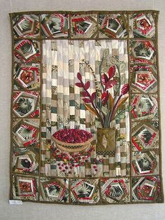 a quilted wall hanging with flowers in a vase and pictures on the wall behind it