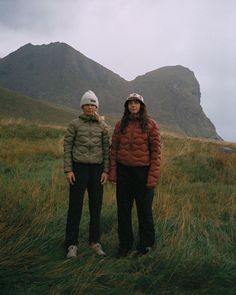 two people standing in the grass with mountains in the background