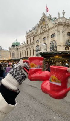 a person holding up two red cups in front of a building with christmas decorations on it