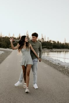 a man and woman holding hands walking down a road next to the water with trees in the background