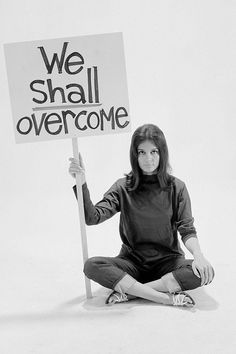 a woman sitting on the ground holding a sign that says we shall overcome with it