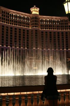 a woman standing in front of a fountain at night with the las vegas hotel and casino behind her