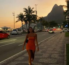 a woman in an orange dress is walking down the street with cars and palm trees