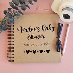 a baby shower book next to a camera, pen and flowers on a pink surface