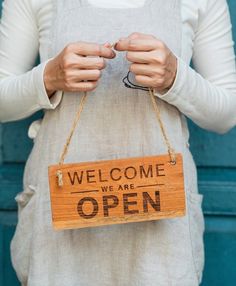 a woman holding a wooden sign that says, welcome we are open on the front