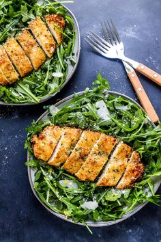 two plates filled with chicken and greens on top of a blue tablecloth next to silverware